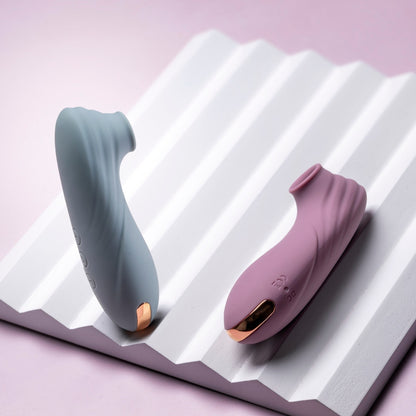 Two clitoral suction vibrators, one teal and one pink, on a white textured surface with a pink background.